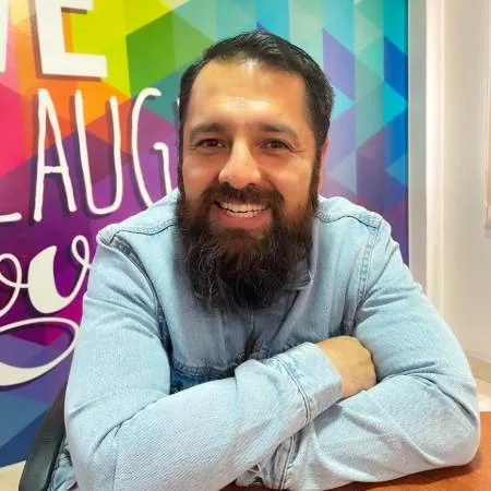 Cesar Vinueza sonriente con barba y camisa de mezclilla sentado en una oficina con fondo colorido.