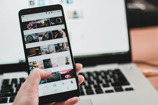 Persona utilizando un teléfono móvil con una galería de fotos abierta, frente a una laptop.